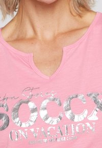 Rosa kortärmad t-shirt med V-ringning. Har metalliskt silvertext som säger "SOCX ON VACATION" och är tillverkad av ett mjukt, texturerat tyg.