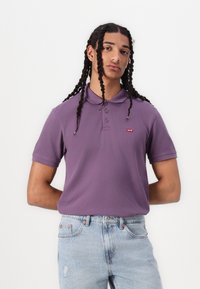Polo shirt viola con colletto, caratterizzato da un piccolo logo rosso Levi's. Abbinato a jeans chiari con strappi. Il modello posa con le mani dietro la schiena.