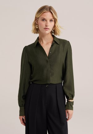 Femme blonde portant des boucles d'oreilles et un bracelet en or, un chemisier vert olive à manches longues et un pantalon noir taille haute sur un fond uni.