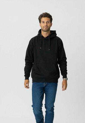 Mand iført en sort hættetrøje og blå jeans, der går fremad med et neutralt ansigtsudtryk mod en ensartet lys baggrund.