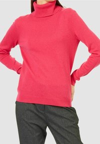 Pull fuchsia avec un col roulé haut et des manches longues, fabriqué en tissu doux. Associé à un pantalon gris, la tenue présente une texture lisse.