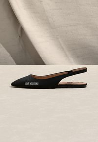 Μαύρο μυτερό φλατ παπούτσι slingback με κείμενο "LOVE MOSCHINO" στο πλάι, ταμπά εσωτερικό πέλμα και μινιμαλιστικό μπεζ υφασμάτινο φόντο.