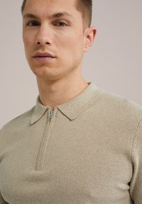 Bežna pletena polo majica z zadrgo in ovratnikom. Tekstura je fina in enakomerna, s subtilnim vzorcem po celi površini.