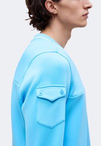 Homme de profil portant un haut bleu clair avec une poche à rabat boutonnée sur la partie supérieure de la manche, debout devant un fond clair et uni.