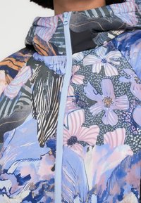 Veste à motif floral mêlant des teintes de bleu, rose et gris, dotée d'une fermeture éclair bleu clair et d'une capuche. Confectionnée en tissu léger.