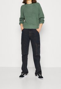 Maglione verde a maglia con una texture a coste e colletto girocollo, abbinato a pantaloni cargo scuri con tasche e sneakers nere.