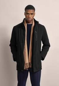 Schwarze, leichte Jacke mit einer Reißverschlusstasche auf der Brust, Knopfverschlüssen und einem hohen Kragen, kombiniert mit einem beigen Schal und einem marineblauen Pullover.