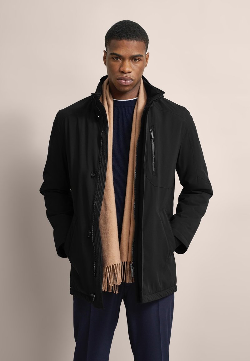 Schwarze, leichte Jacke mit einer Reißverschlusstasche auf der Brust, Knopfverschlüssen und einem hohen Kragen, kombiniert mit einem beigen Schal und einem marineblauen Pullover.