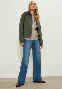 Oliven grøn, letvægtsjakke med en mørk lilla krave, parret med en beige kabelstriktrøje og brede blå jeans. Brune ankelstøvler.