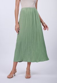Svetlozelená plisovaná maxi sukňa z textúrovanej tkaniny, s elegantne plynúcim pásom. Nosí sa s béžovými sandálmi na podpätku.