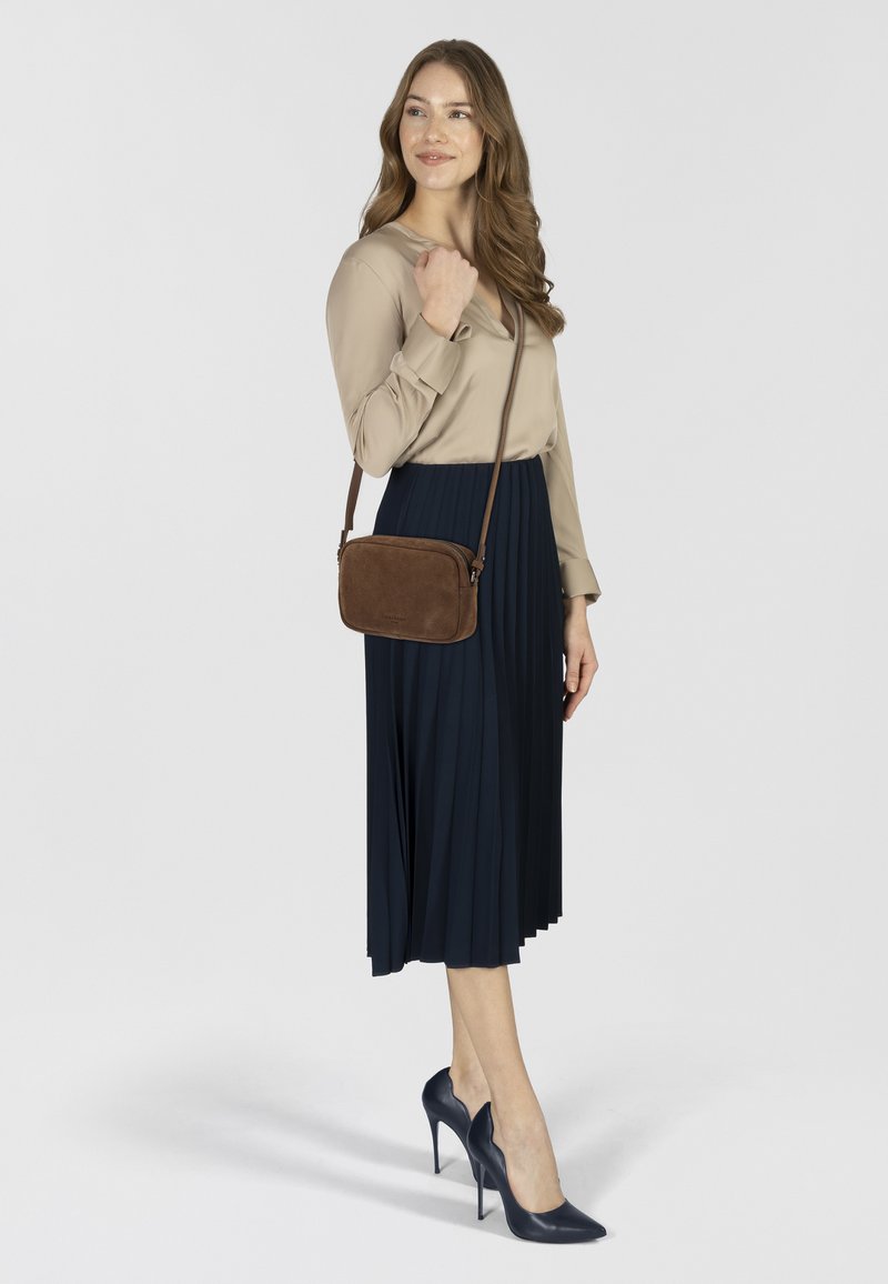 Blusa beige, gonna a pieghe blu scuro, borsa a tracolla in suede marrone e tacchi alti blu scuro. Il modello sta in piedi con la mano destra sulla borsa, girato leggermente a destra.