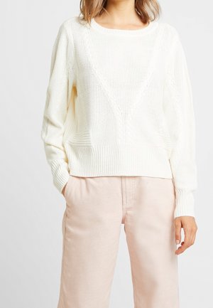 Personne portant un pull en laine blanc à col rond et un pantalon beige clair, les mains dans les poches.