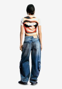 Orezané biele tričko s grafikou úst; modré nadmerné džínsové cargo nohavice s vreckami a vyblednutým vzhľadom, nosené s čiernymi topánkami.