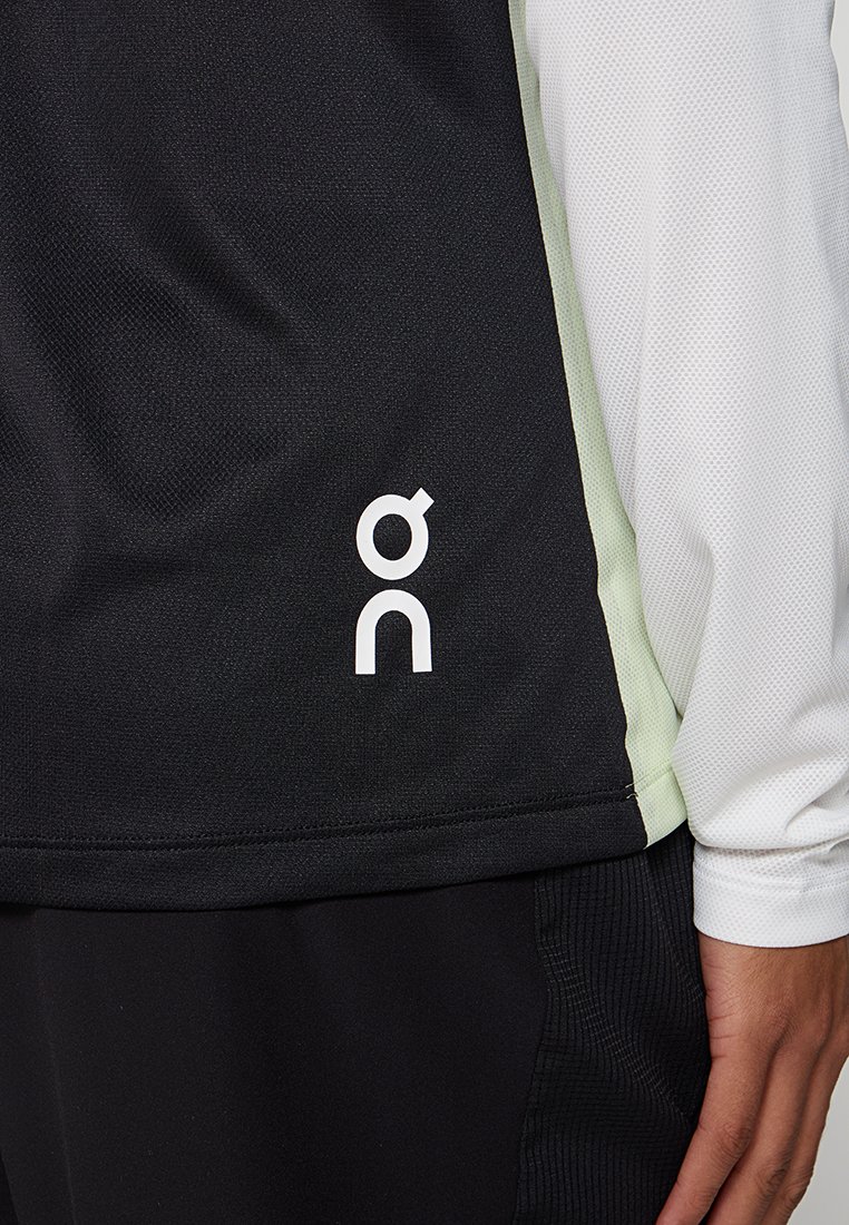 Czarny i jasnozielony sportowy top z białym logo po boku, noszony z czarnymi spodniami i białą długą koszulką pod spodem.