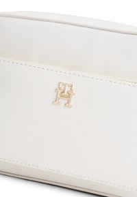 Λευκή δερμάτινη τσάντα με διακοσμητικό χρυσό λογότυπο. Λείο υλικό με εμφανείς ραφές και κομψό, λιτό σχέδιο. Κλείσιμο με φερμουάρ στην κορυφή.