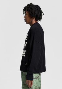 Maglione a maglia nera con testo bianco, polsini e orlo a coste, vestibilità rilassata. Il modello indossa pantaloni verdi, evidenziando il dettaglio della catena in vita.