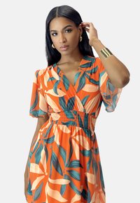 Robe florale orange avec des motifs de feuilles, manches courtes et taille smockée. Le tissu est léger et semi-transparent. Bracelet manchette en or en accessoire.