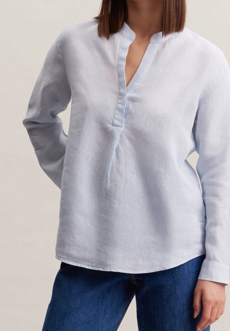 Femme portant un chemisier en lin bleu clair à manches longues avec une encolure fendue et un jean bleu, debout avec une main dans le dos.