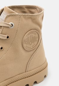 Botte montante en toile beige avec écusson rond en caoutchouc portant l'inscription « Palladium L'Originale Depuis 1947 » et semelle en caoutchouc texturée.