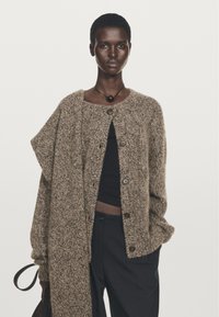 Cardigan lavorato a maglia di colore marrone chiaro con un design testurizzato, caratterizzato da grandi bottoni e uno stile aperto. Indossato sopra un top nero.