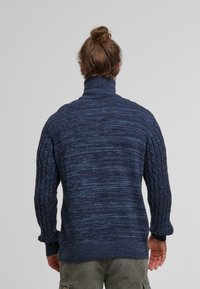 Granatowy sweter dzianinowy z wysokim kołnierzem, z teksturowanym wzorem w warkocze na rękawach oraz gładką częścią główną. Regulowane mankiety.