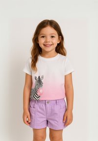 Fehér póló zebra grafikával és rágógumival, fokozatos rózsaszín színben; világos lila rövidnadrággal párosítva, zsebekkel és patentos záródással.
