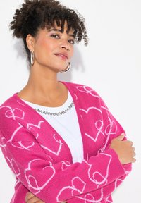 Růžový pletený cardigan s bílými vzory srdcí, širokými rukávy a výstřihem do V. V páru s bílou košilí s výstřihem zdobeným kamínky.