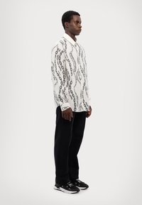 Camisa blanca de manga larga con patrón de cadena negra, cuello abotonado y pantalones negros. El modelo lleva zapatillas blancas y negras.