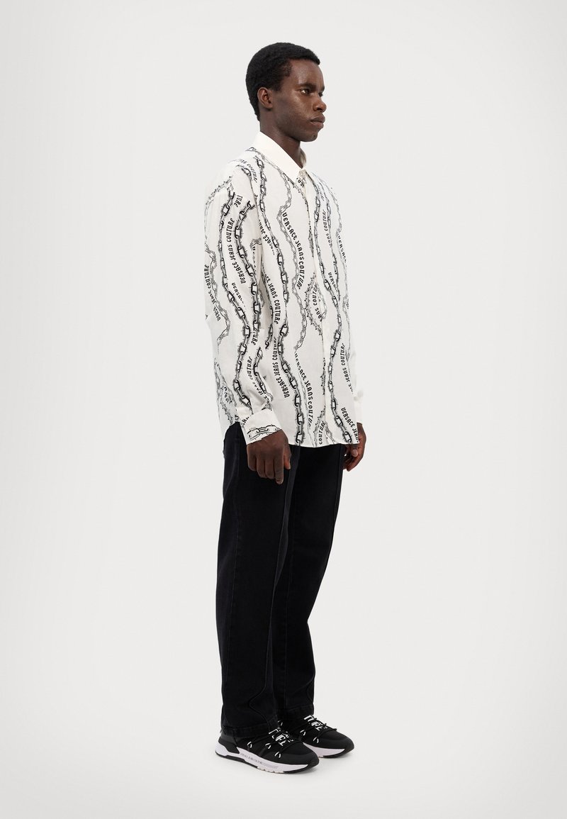 Camisa blanca de manga larga con patrón de cadena negra, cuello abotonado y pantalones negros. El modelo lleva zapatillas blancas y negras.