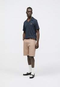 Tengerészkék rövid ujjú galléros póló sárga logóval, bézs csíkos rövidnadrággal és fekete cipővel, fehér zoknival. Kényelmes, hétköznapi öltözet.