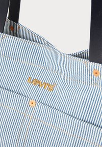 Striped denim tote bag in blue and white with orange stitching and a gold "Levi's" logo. Features durable black handles and button accents.