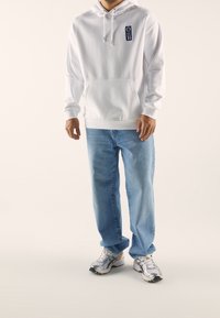 Felpa bianca con cappuccio, tasca frontale, cordino e toppa blu; abbinata a jeans chiari a vestibilità ampia e sneakers argentate.