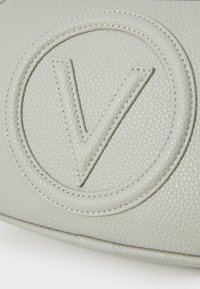 Ανοιχτό γκρίζο δερμάτινο τσάντα με έντονο ανάγλυφο λογότυπο. Η υφή είναι pebbled με αντίθετα ράμματα που περιγράφουν το σχέδιο.