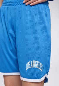 Pantaloni sportivi in rete blu con bordi bianchi, con la scritta "LOS ANGELES" stampata in bianco in testo arcuato sulla parte bassa della gamba.
