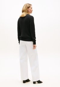 Schwarzer, gestrickter Pullover mit einer strukturierten Musterung, kombiniert mit locker sitzenden weißen Hosen. Das Modell steht mit dem Rücken zur Kamera.