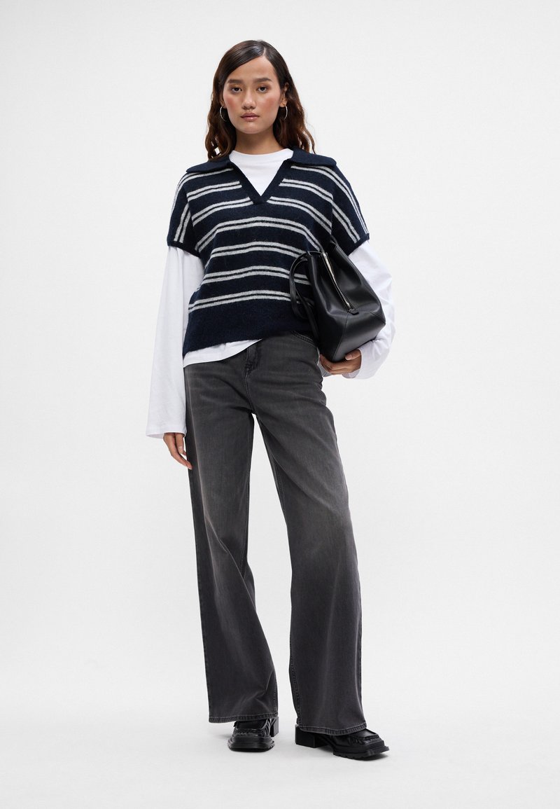 Niebiesko-białe w paski dzianinowe kamizelka z dekoltem w serek na białej koszuli z długim rękawem, zestawiona z szarymi szerokimi spodniami i czarną torebką.