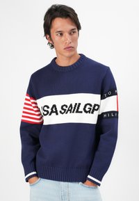 Young man wearing a navy blue sweater with white, red stripes, and "USA SAIL GP" text, standing with hands in light blue jeans pockets.