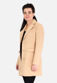 Abrigo de mezcla de lana beige con cuello con muescas, dos bolsillos frontales, tres botones y corte recto, exhibido en un modelo sobre un fondo blanco.