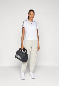 Weißes T-Shirt mit kurzen Ärmeln und schwarzen Streifen, beigefarbene Jogginghose und schwarze Adidas Reisetasche mit weißem Logo. Glattes Material, lockere Passform.
