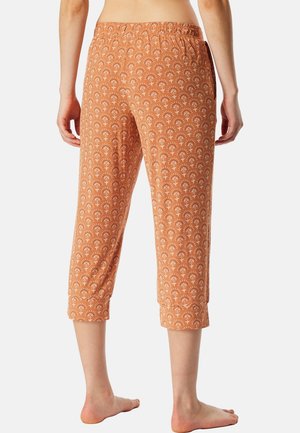 Personne de la taille vers le bas portant un pantalon orange mi-mollet à taille élastique avec un petit motif floral blanc, debout pieds nus sur un fond uni.
