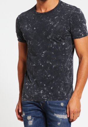 Ein Mann trägt ein schwarz-grau marmoriertes kurzärmeliges T-Shirt und abgenutzte blaue Jeans und steht vor einem schlichten Hintergrund.
