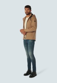 Licht beige jas met een capuchon, met een zwarte voering en rits, gedragen over een crèmekleurig shirt, gecombineerd met blauwe jeans en zwarte laarzen.