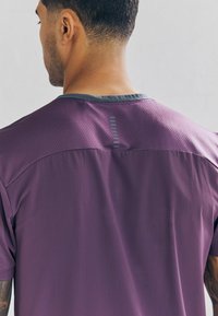 Paarse sportshirt van textuurstof, met een bovenkant van mesh en een grijze halslijn. Heeft een verticale streepdetail op de achterkant.