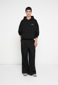 Hanorac negru cu glugă și buzunar frontal, combinat cu pantaloni evazați cu carouri în tonuri închise. Încălțămintea are un design stratificat, în două tonuri.