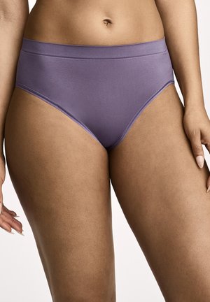 Frau trägt hoch taillierte, glatte lila Unterwäsche und zeigt Bauch- und oberen Oberschenkelbereich vor weißem Hintergrund.