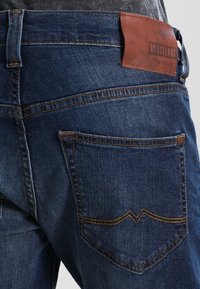 Vaqueros de mezclilla azul oscuro con una etiqueta de cuero etiquetada "Mustang" y costuras amarillas en contraste. El bolsillo trasero tiene un patrón de costura distintivo.