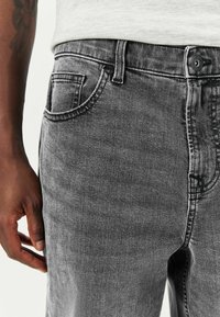 Hallid hallid teksad, millel on sile tekstuur, viie tasku disain, vöökohaga ja lukuga sulgemine. Suumvaade puusa ala.