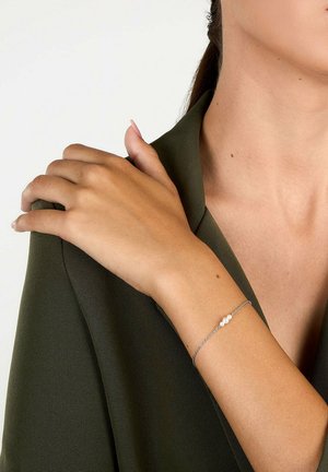 Frau trägt dunkelgrünen Blazer, berührt die Schulter und zeigt filigranes silbernes Kettenarmband mit drei kleinen weißen Perlen.