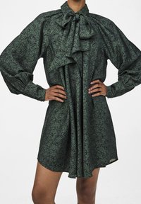 Green floral patterned dress with a high neck, large bow, and long puff sleeves; made of lightweight fabric, pleated at the front.