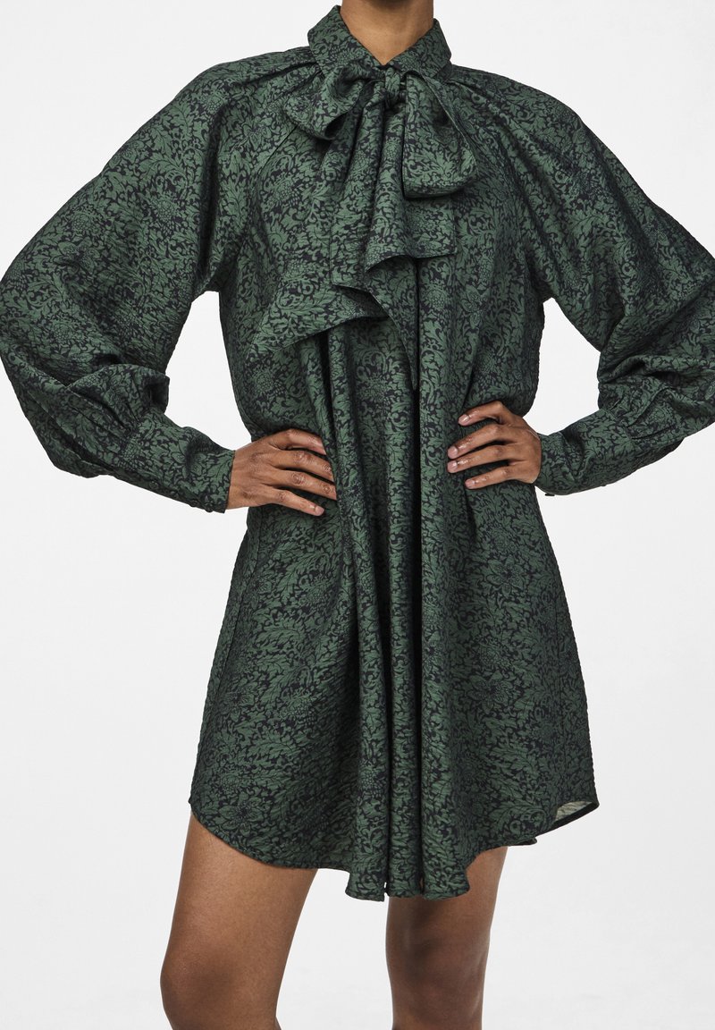Green floral patterned dress with a high neck, large bow, and long puff sleeves; made of lightweight fabric, pleated at the front.